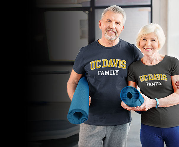 An aged couple is wearing UC DAVIS family tees and holding a yoga carpet.
